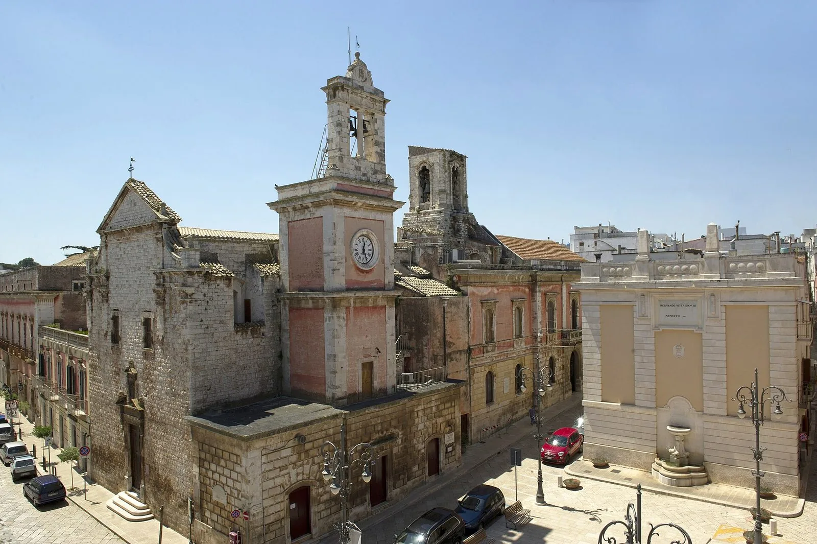 Il centro di Castellana Grotte visto dall'alto in una giornata di sole: la facciata in pietra della chiesa di San Francesco, la torre dell'orologio rosata con il campanile, il palazzo municipale e una piazza lastricata con lampioni in ferro battuto