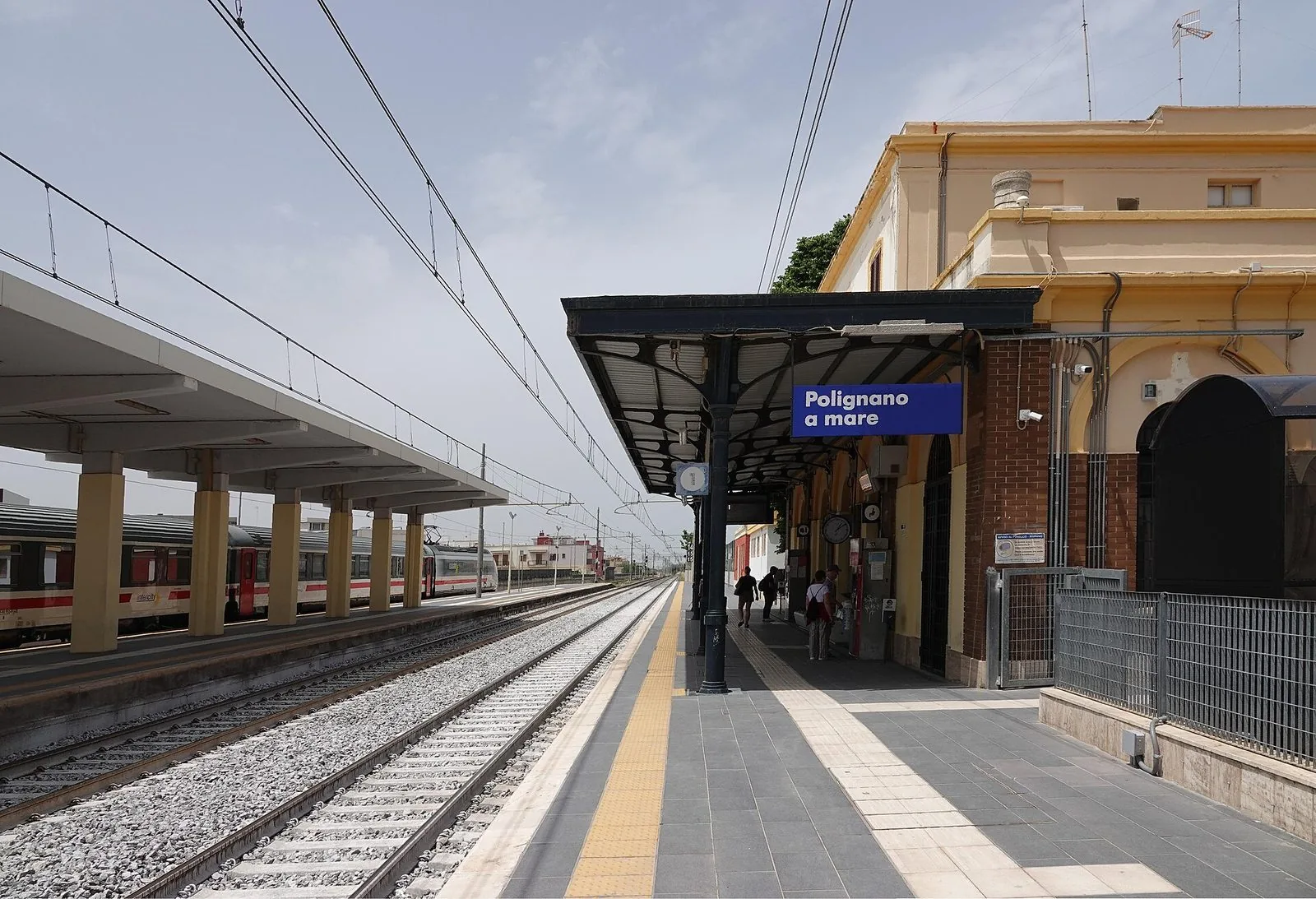 Il binario della stazione di Polignano a Mare con la pensilina in ferro, il cartello blu con il nome della città, l'edificio color crema e un treno fermo sul binario accanto