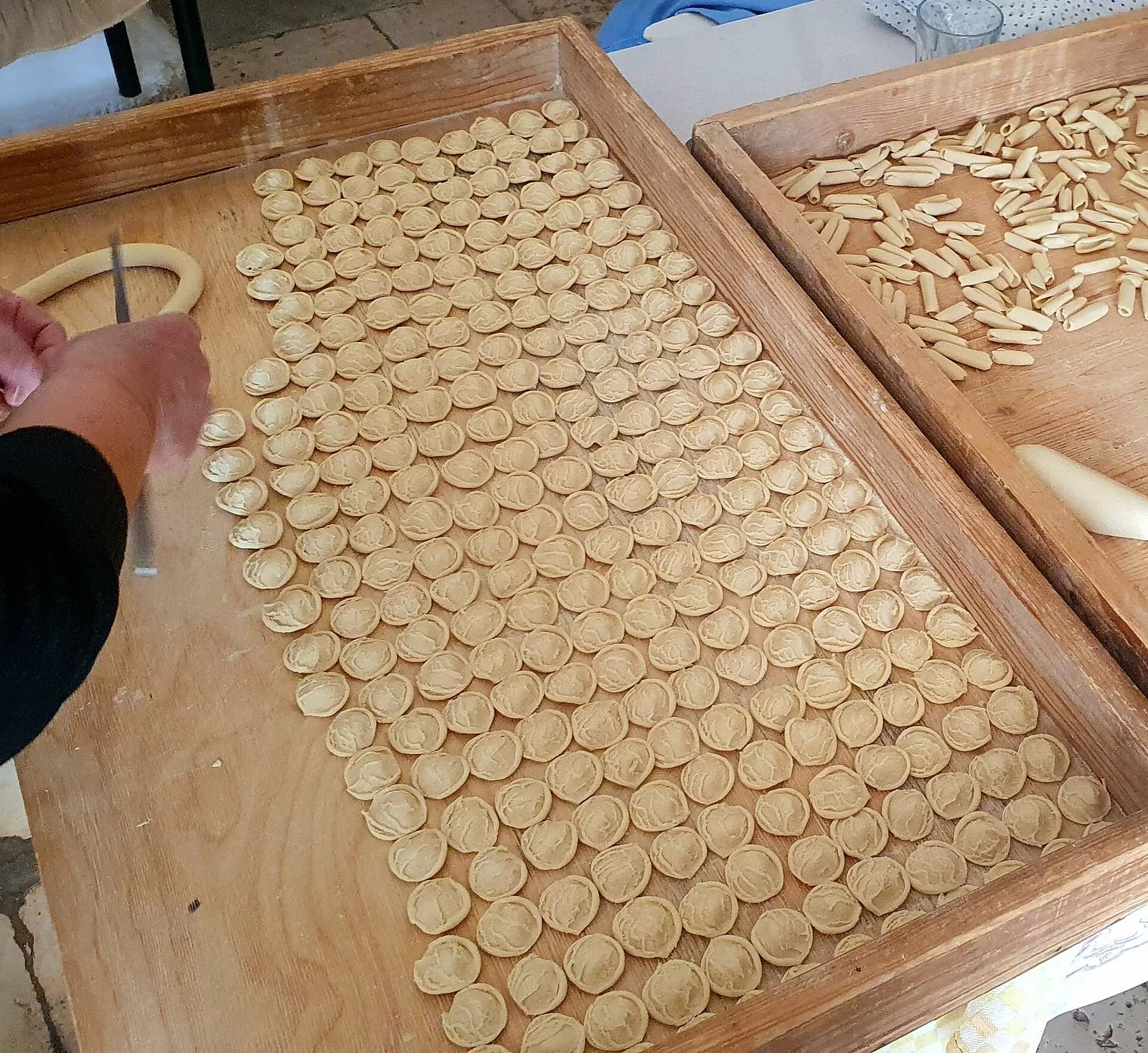 Orecchiette fatte a mano disposte in file ordinate su una grande spianatoia di legno, con accanto un vassoio di cavatelli e una mano che lavora l'impasto con il coltello