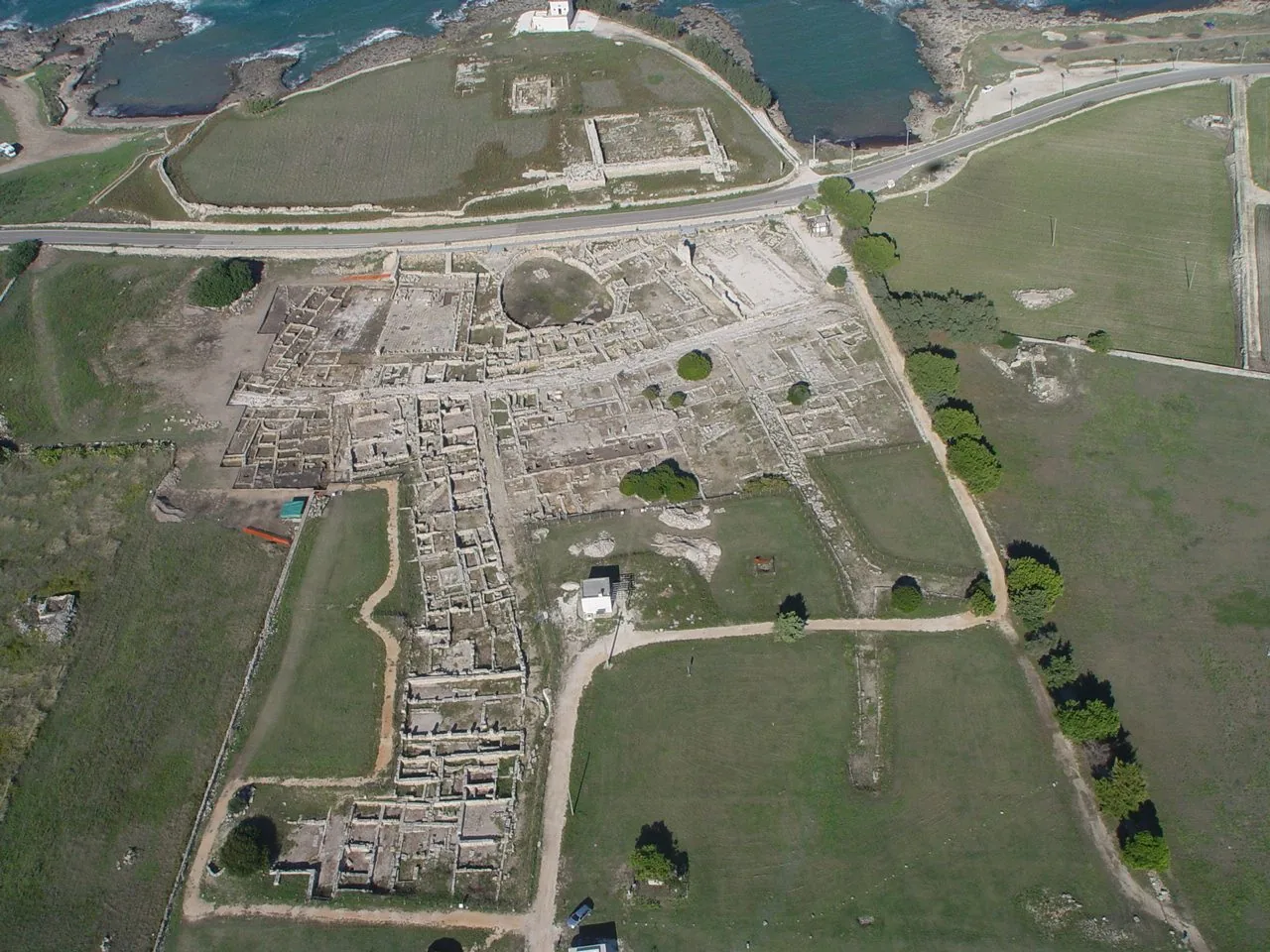 Veduta aerea del Parco archeologico di Egnazia: i resti in pietra della città antica tra prati verdi, la strada costiera e, in alto, l'acropoli affacciata sul mare con le insenature rocciose