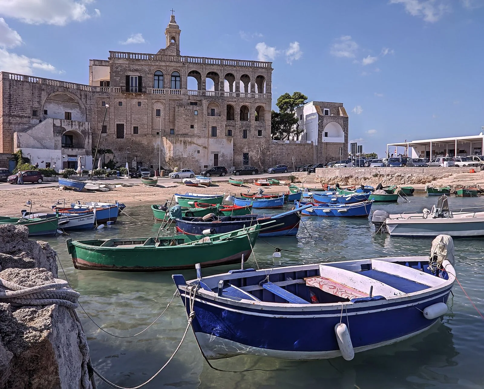 L'Abbazia di San Vito a Polignano a Mare con la facciata a loggiati e il campanile, affacciata sul piccolo porto pieno di barche da pesca blu, verdi e bianche nell'acqua limpida