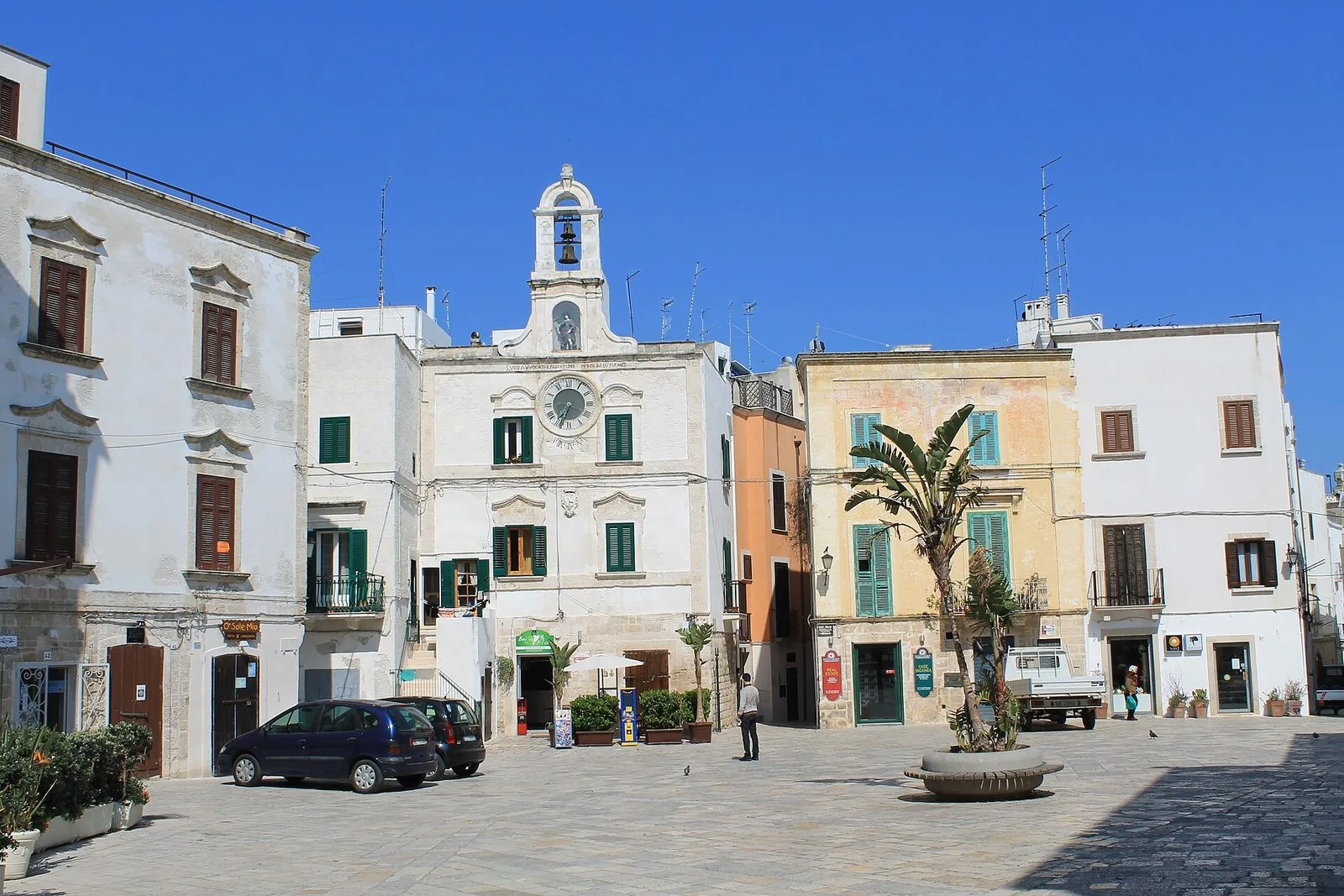 Piazza dell'Orologio nel centro storico di Polignano a Mare: il palazzo bianco con orologio e campanile a vela con la statua del santo, case dalle persiane verdi e una palma al centro della piazza lastricata