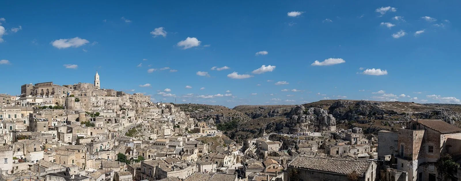 Panorama dei Sassi di Matera sotto un cielo azzurro con nuvole bianche: a sinistra le case in pietra chiara che salgono verso il campanile della Cattedrale sulla Civita, a destra la gravina e l'altopiano roccioso della Murgia