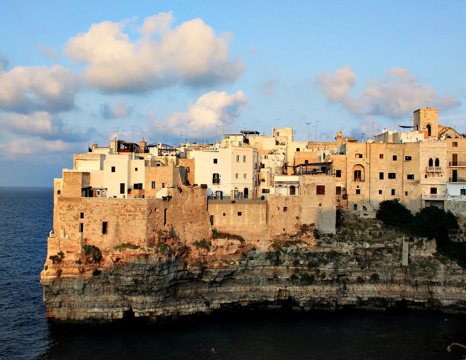 Il centro storico di Polignano a Mare affacciato sul mare, con case bianche e color pietra costruite sul bordo della scogliera, illuminate dalla luce calda del tardo pomeriggio sotto un cielo con nuvole bianche