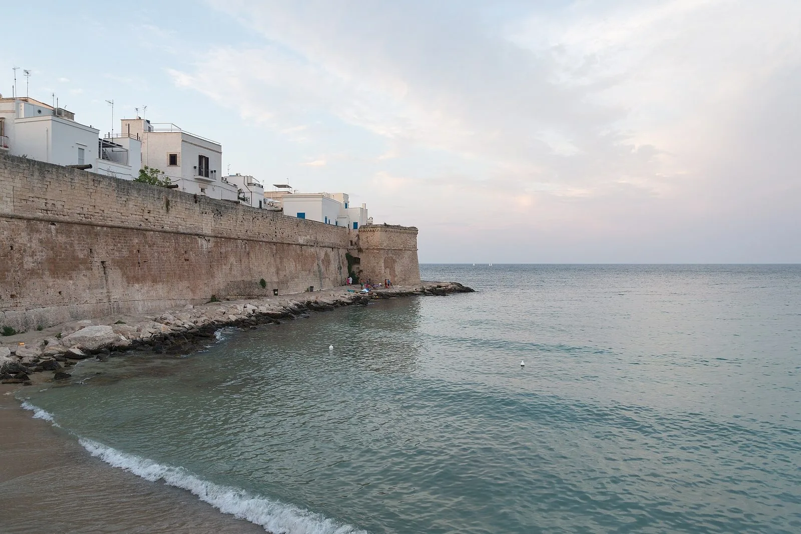 Cala Porta Vecchia a Monopoli: una piccola spiaggia di sabbia lambita dall'onda, ai piedi delle antiche mura in pietra con un bastione e le case bianche affacciate sul mare calmo