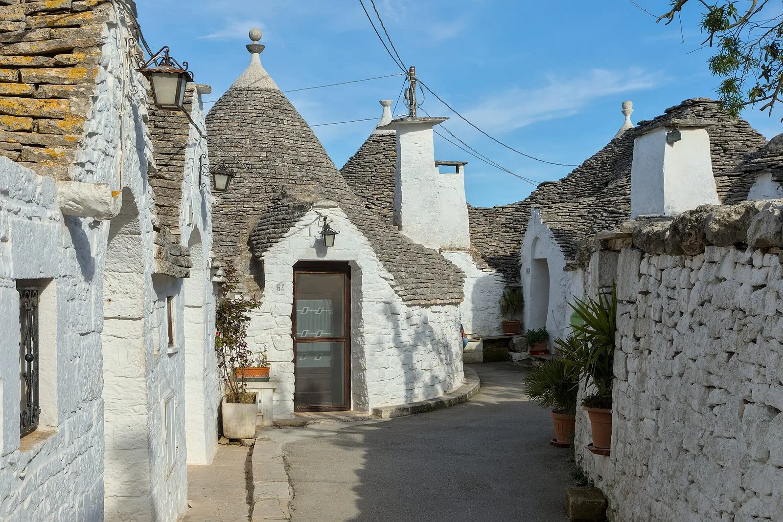 Un vicolo del Rione Monti ad Alberobello: trulli imbiancati a calce con i tetti conici di pietra grigia e i pinnacoli, lanterne in ferro, vasi di piante e un muro in pietra bianca sotto il cielo azzurro