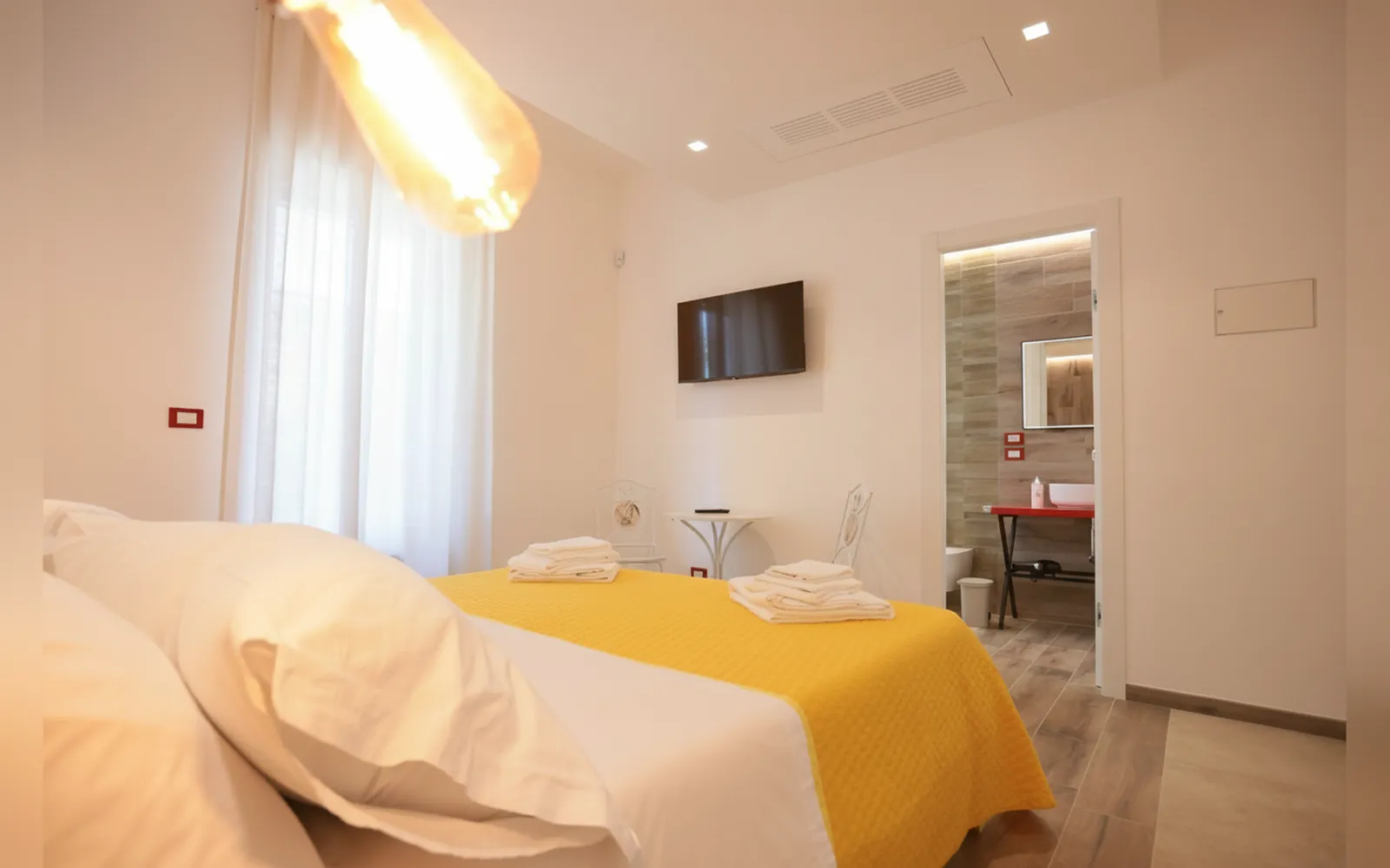 Il letto con il copriletto giallo, la TV a parete e la porta del bagno